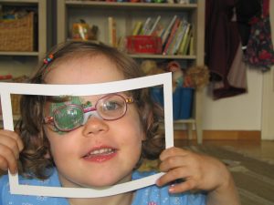 Frühförderung für Kinder mit Sehschädigung – Kind mit Occlusionspflaster auf Auge schaut durch gebastelten Papier-Bilderrahmen